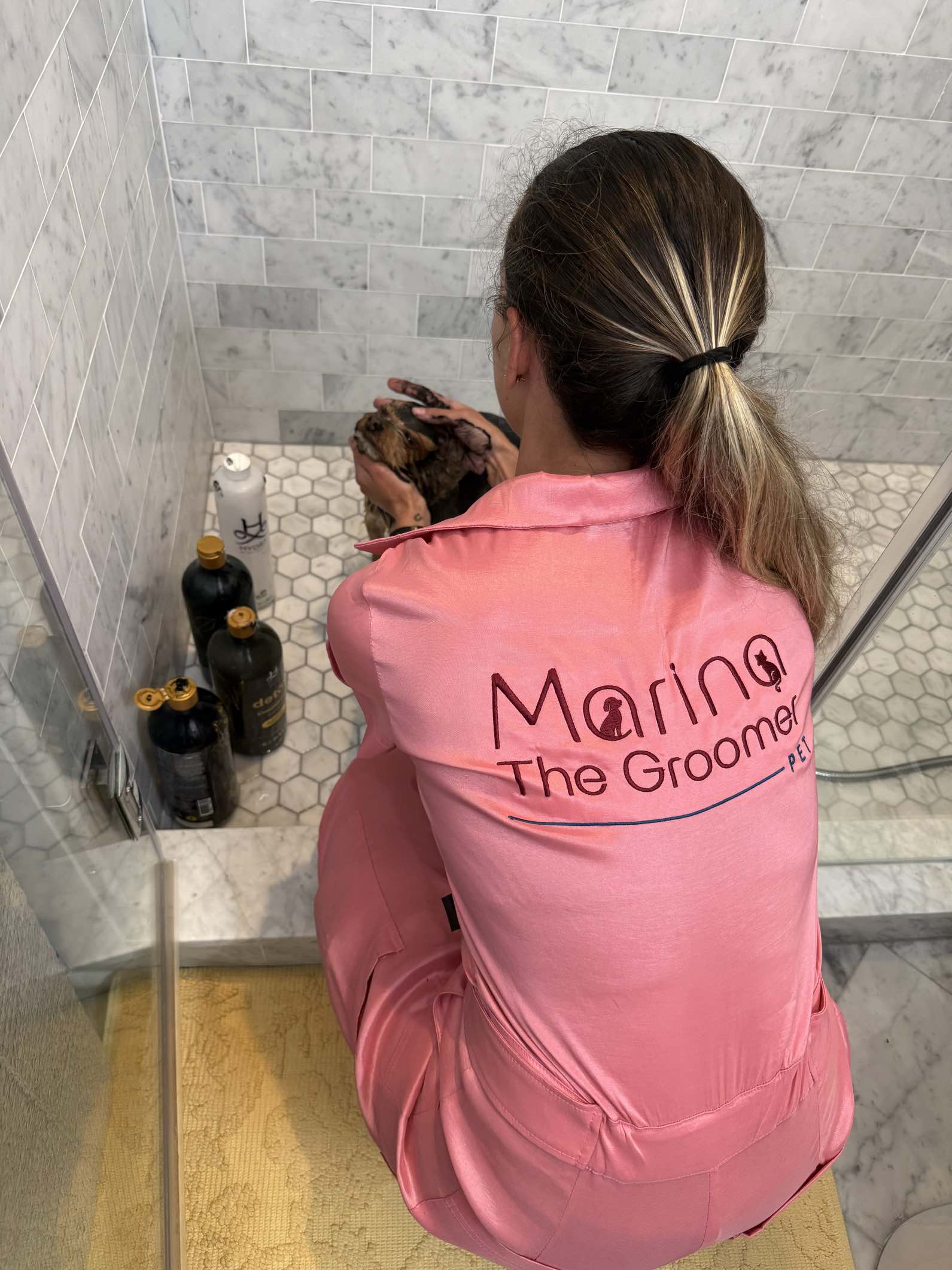 Marina in her pink uniform gently bathing a small dog in a marble house-call grooming setup