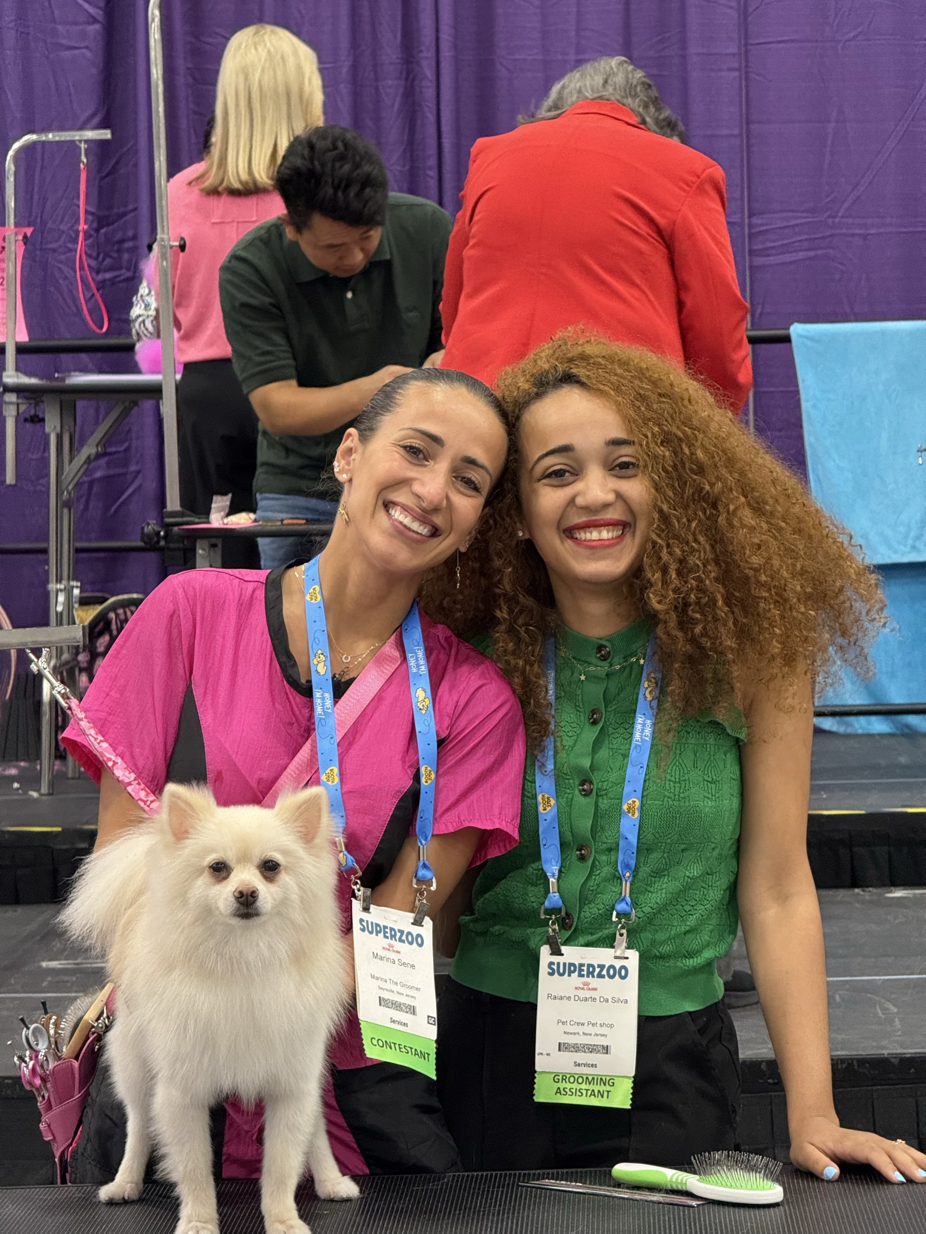 Raiane and Marina smiling together with a fluffy white Pomeranian at a grooming event